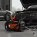 Een robuuste, oranje en zwarte jumpstarter met klemmen en kabels ligt op de vloer van een garage, met een open motorkap van een auto op de achtergrond.