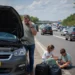 Automobilist belt pechhulp naast een auto met open motorkap langs een drukke snelweg.